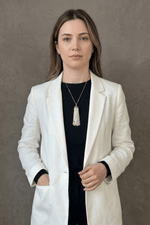 Kadın, beyaz ceket giymiş, püsküllü doğal inci kolye takıyor. Arka plan düz gri.