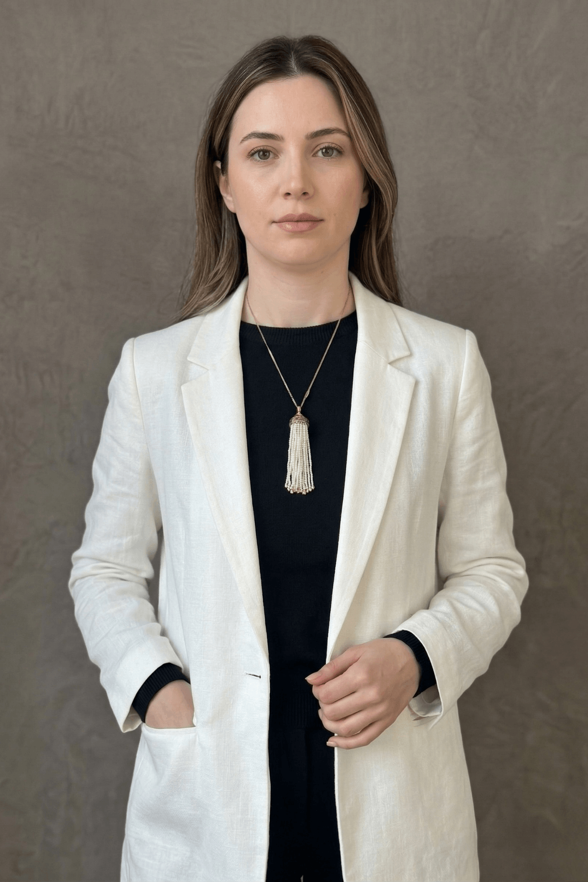 Kadın, beyaz ceket giymiş, püsküllü doğal inci kolye takıyor. Arka plan düz gri.